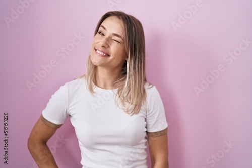 Blonde caucasian woman standing over pink background winking looking at the camera with sexy expression, cheerful and happy face.