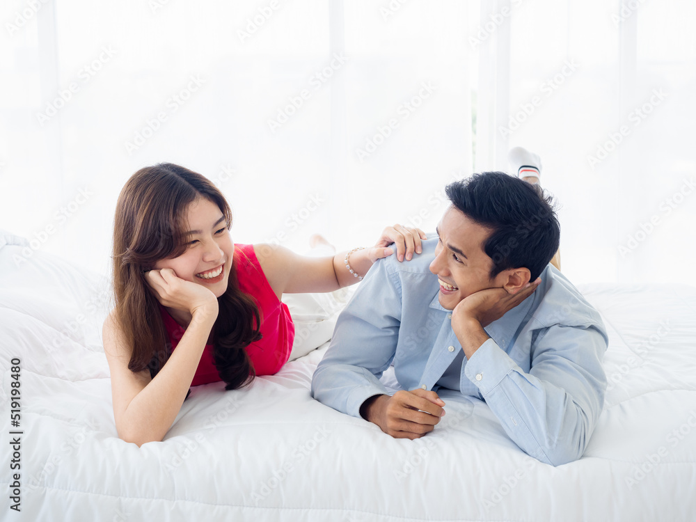 The sweet moment of happy couple lying on the white bed. Asian man in denim shirt and young beautiful woman smile and talk together on bed in white bedroom. Honeymoon trip on summer vacation.