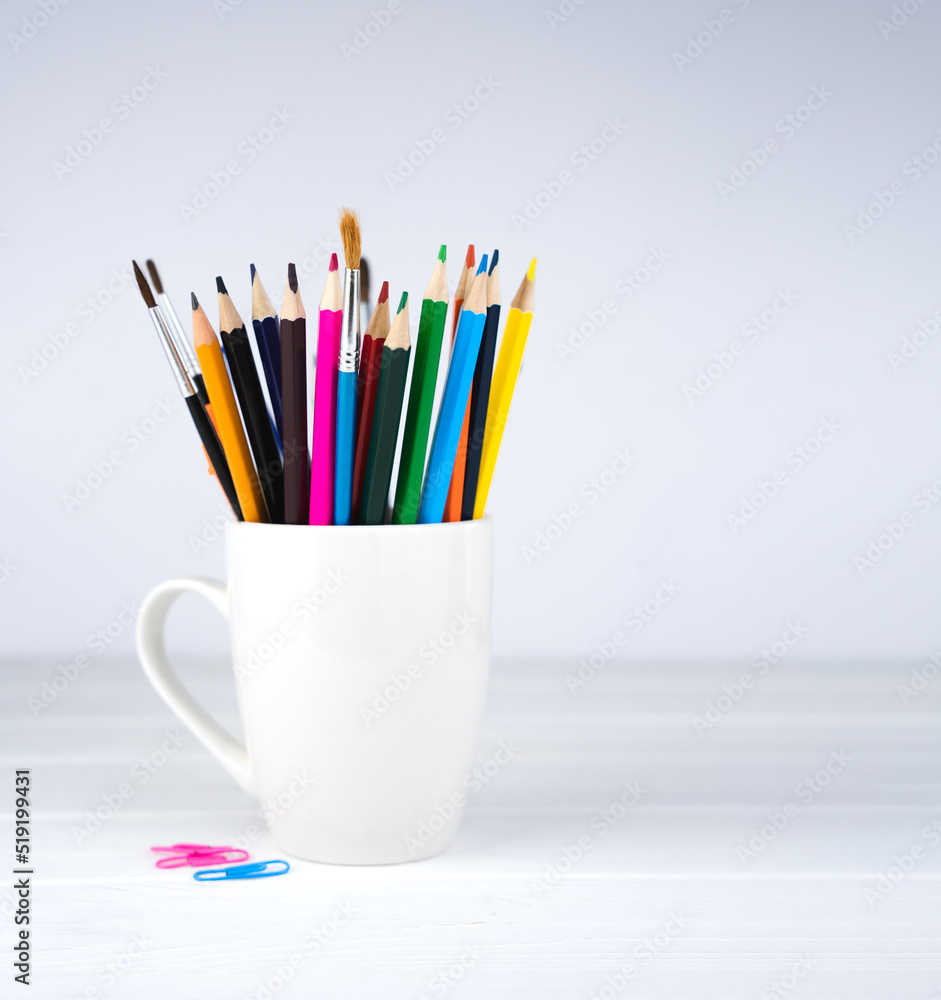 Pencils and brushes in a white mug on a white wooden background. Minimalist composition. Place for text. Selective focus. Pencils and brushes in a white mug on a white wooden background. Minimalist composition. Place for text. Selective focus.