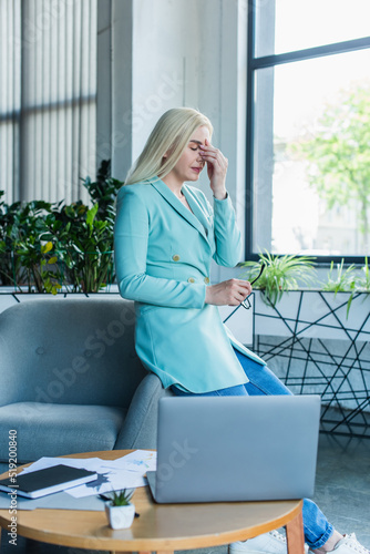 Tired psychologist holding eyeglasses near laptop in consultation room.