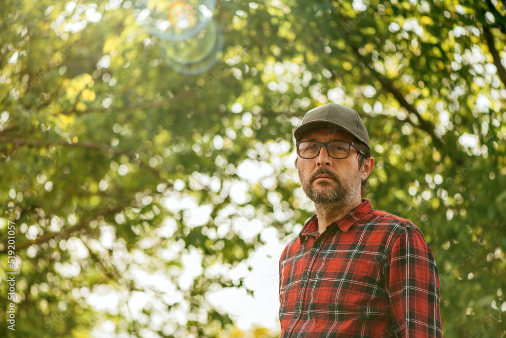 Obraz premium Portrait of male farmer wearing plaid shirt and trucker's hat posing in walnut orchard and looking at camera