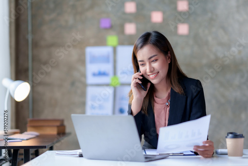 Charming and smiling Asian businesswoman chatting on mobile phone with customer in office.