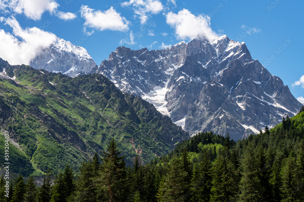 Beautiful alpian mountains landscape. High snow covered mountains green high forset on the hills under blue sky with clouds.