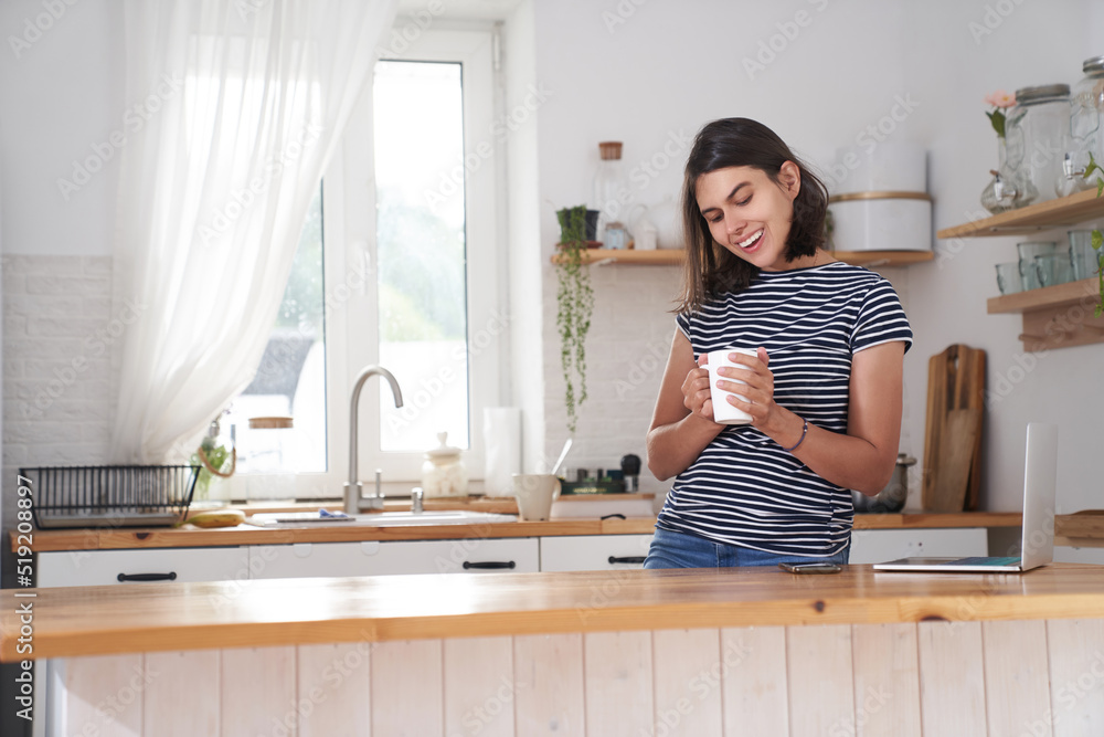 A woman in the kitchen with a smile. A woman drinks coffee and holds a cup in her hand. She is happy because She drinks her favourite coffee in the morning. High quality photo with copy space.