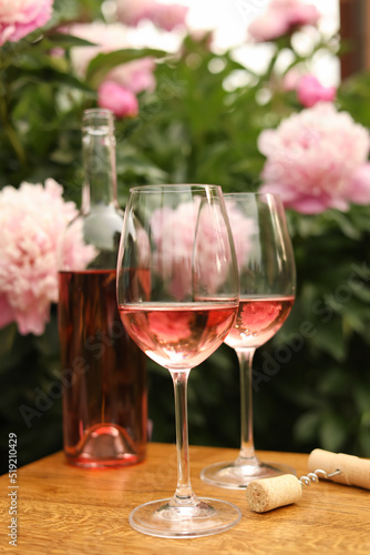 Glasses and bottle with rose wine on wooden table near beautiful peonies