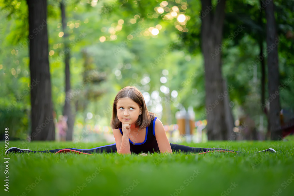 little girl doing yoga sits on the lawn in the park in a twine and looks at the camera