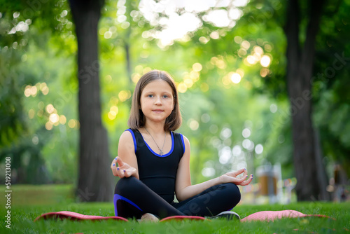 a little girl sitting in a park meditates in a folded lotus position and looks at the camera © Roman
