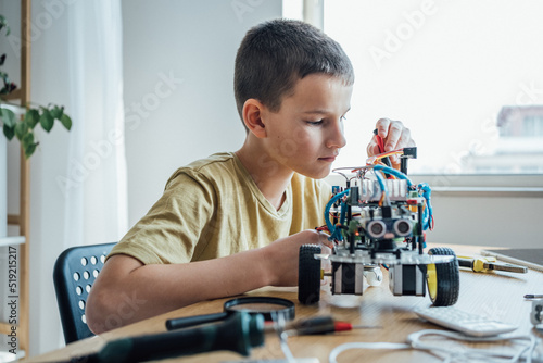 Small nerd is holding a robot. Boy is sitting at a table in front of robot car. He is very satisfied with his work and feeling concentrated