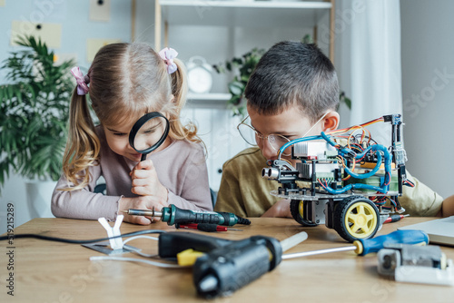 Happy young girl looking through the magnifying glass with her brother while building electronic robot together. Education, science and children concept