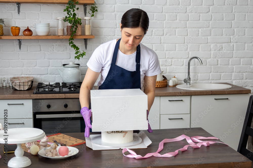 Woman in an apron covers cake with box for packing for delivery. Selective focus. Photos about confectioners, food, hobbies. Woman in an apron covers cake with box for packing for delivery. Selective focus. Photos about confectioners, food, hobbies.