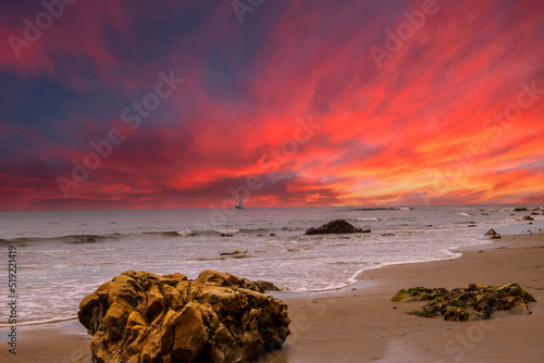 a sail boat sailing across the vast blue ocean water with large rocks in the silky brown sands of the beach with powerful clouds at sunset at Leadbetter Beach in Santa Barbara California USA photo