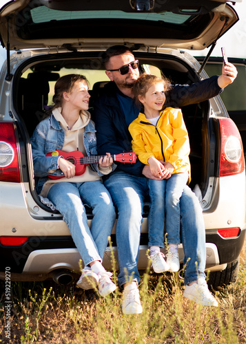 A dad with two daughters in the car. They sit in the open trunk of the car. The girl plays the ukulele. Dad takes a selfie. Communication. Family vacation.