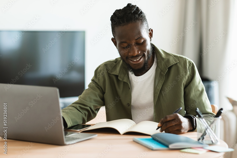 Smiling african american man sitting at desk, working on laptop and taking notes in notebook, black male studying online Smiling african american man sitting at desk, working on laptop and taking notes in notebook, black male studying online
