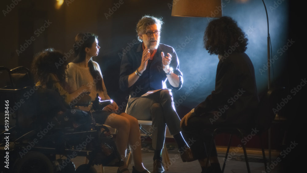 A group of actors with a disabled actress and a director, with a script sitting on a dark stage and discussing a theater performance together.