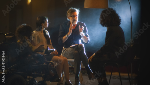 A group of actors with a disabled actress and a director, with a script sitting on a dark stage and discussing a theater performance together.