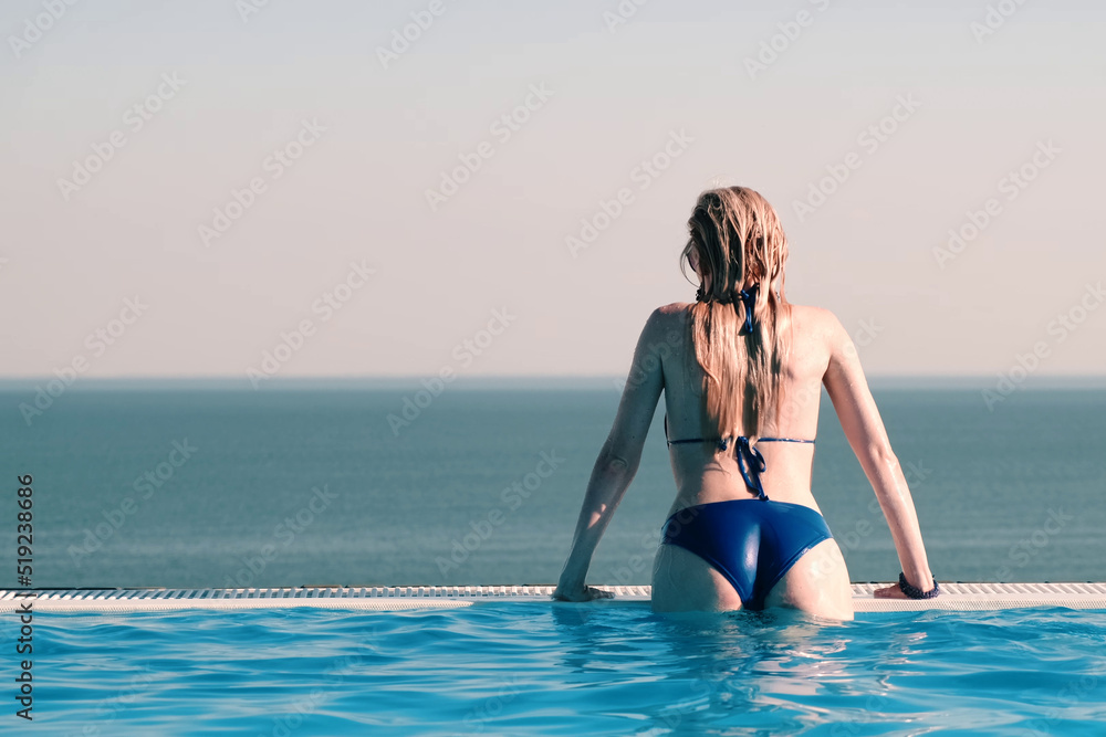Beautiful girl in a blue swimsuit enjoys relaxing in the infinity pool overlooking the sea. Rising out of the water rests on the edge of the pool. Back view