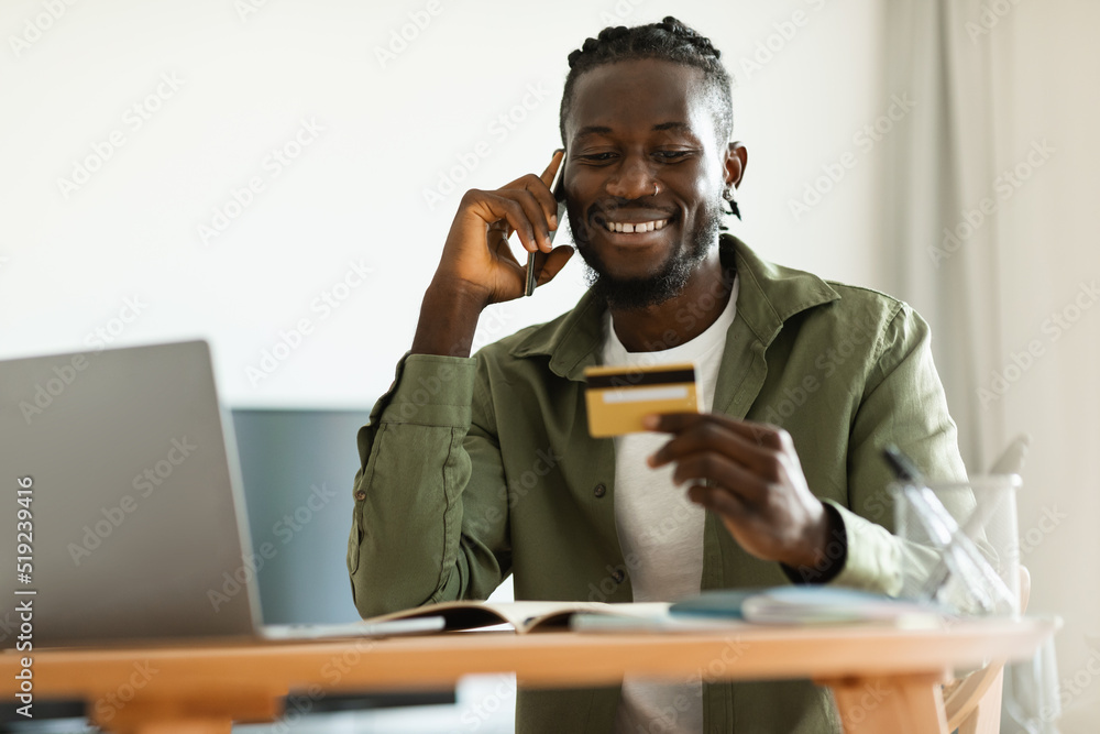 Excited black man holding credit card, having phone conversation with bank representative, sitting at home Excited black man holding credit card, having phone conversation with bank representative, sitting at home