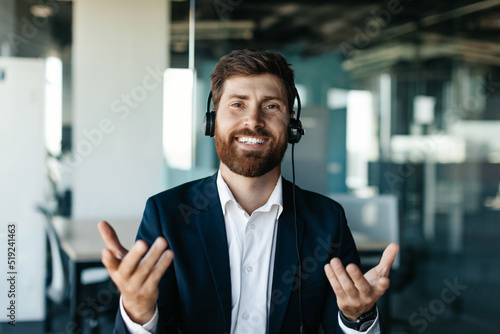 Portrait of happy businessman in headset talking and gesturing at camera, sitting in office interior, copy space photo
