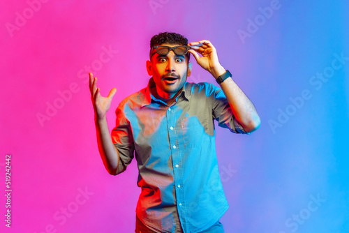 Portrait of surprised man in shirt standing raised glasses and hand, looking at camera with big eyes and shocked expression. Indoor studio shot isolated on colorful neon light background.