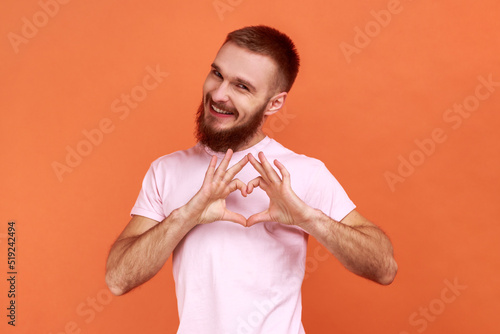 Portrait of smiling bearded man standing with heart or love gesture and looking at camera with toothy smile, wearing pink T-shirt. Indoor studio shot isolated on orange background.