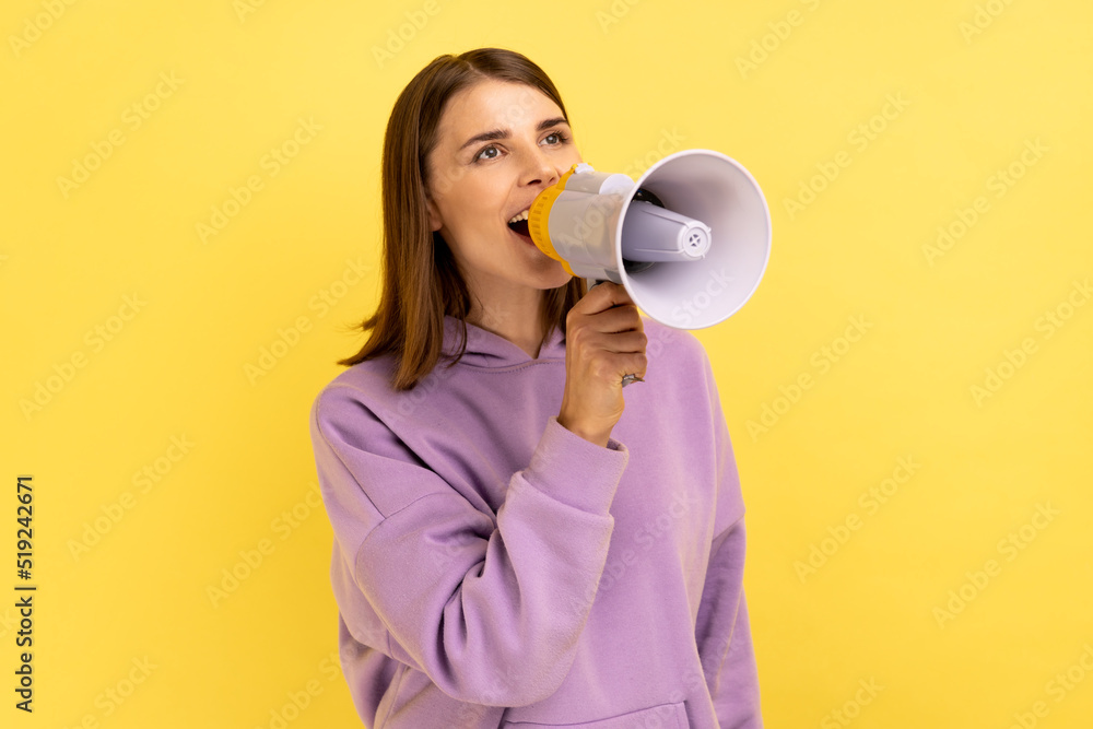Portrait of beautiful woman screaming loudly in megaphone, announcing important advertisement, protesting, wearing purple hoodie. Indoor studio shot isolated on yellow background. Portrait of beautiful woman screaming loudly in megaphone, announcing important advertisement, protesting, wearing purple hoodie. Indoor studio shot isolated on yellow background.