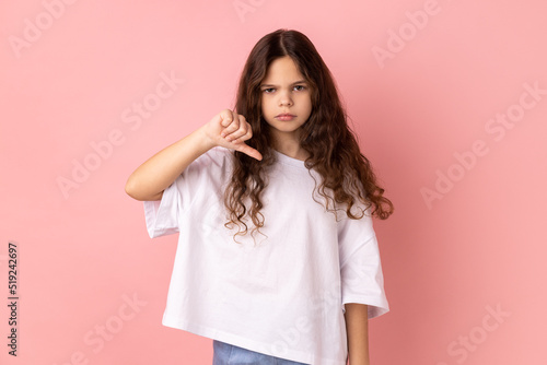 Portrait of little girl in white T-shirt criticizing bad quality with thumbs down displeased grimace, showing dislike gesture, expressing disapproval. Indoor studio shot isolated on pink background.