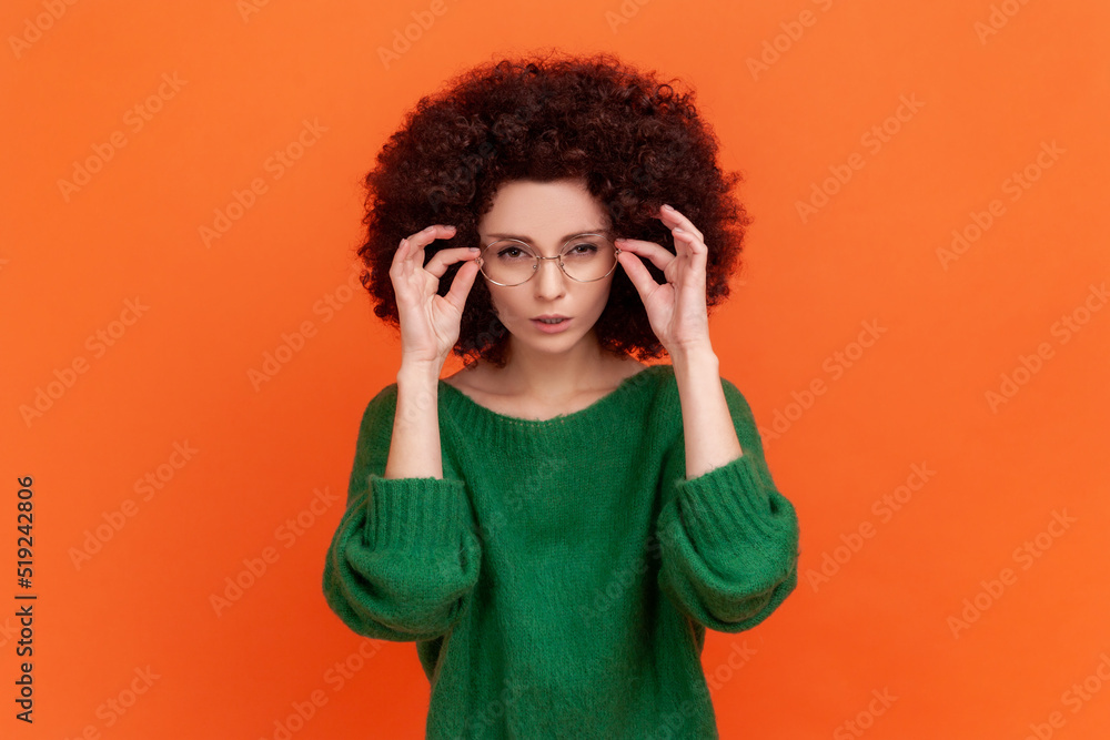 Portrait of attentive beautiful woman with Afro hairstyle wearing green casual style sweater standing holding glasses and looking at camera. Indoor studio shot isolated on orange background. Portrait of attentive beautiful woman with Afro hairstyle wearing green casual style sweater standing holding glasses and looking at camera. Indoor studio shot isolated on orange background.