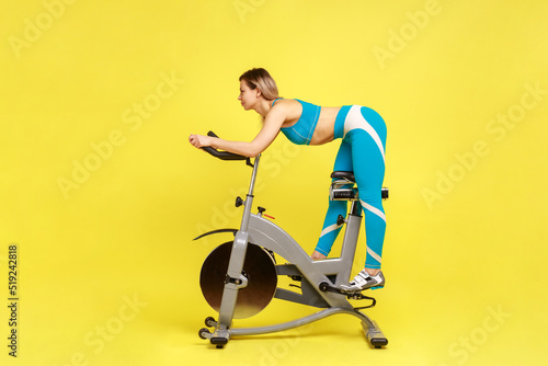 Side view of slim attractive sporty woman on exercise bike, looking ahead with calm expression, enjoying workout, wearing blue sportswear. Indoor studio shot isolated on yellow background.