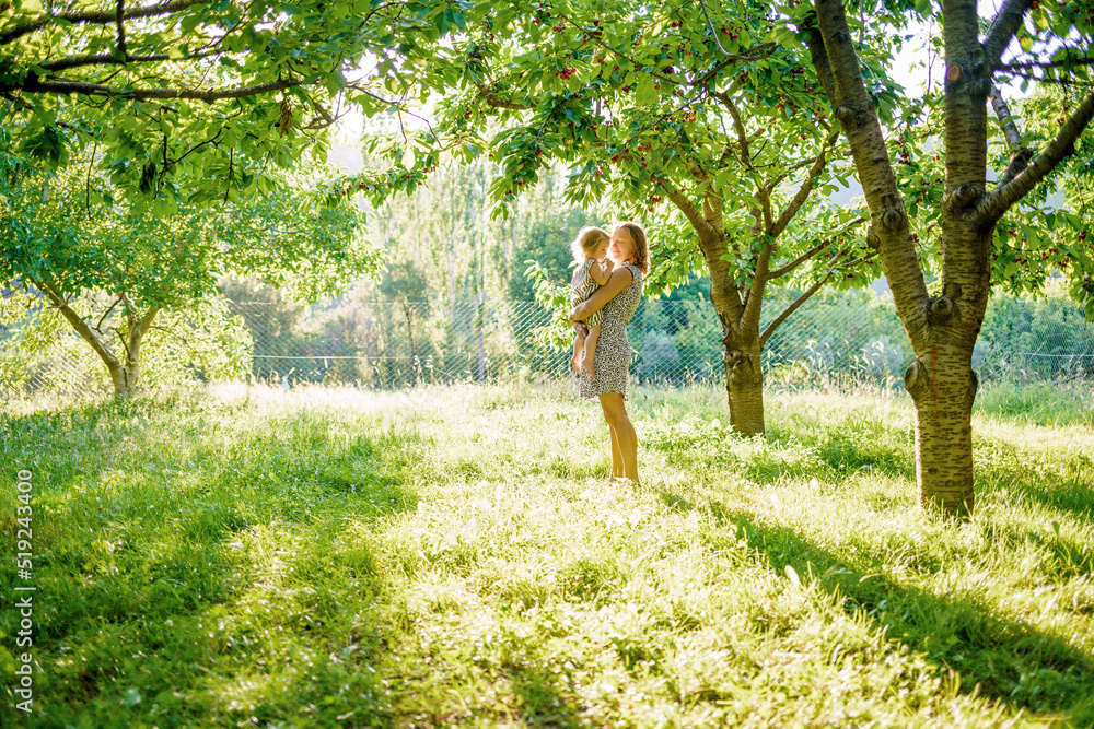 The joy of motherhood. Mom and daughter in the cherry orchard. Sunbeams on a green meadow The joy of motherhood. Mom and daughter in the cherry orchard. Sunbeams on a green meadow