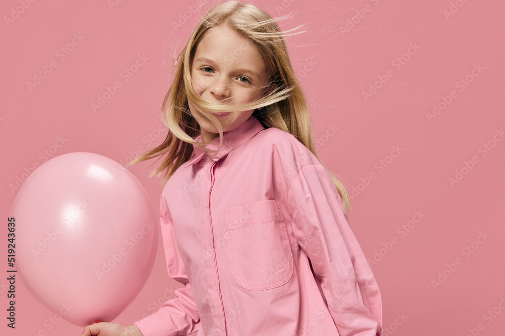 a beautiful, happy school-age girl stands with her hair flying in the wind holding a large pink balloon in her hand in a pink dress on a pink background with a blank space for an advertising mockup a beautiful, happy school-age girl stands with her hair flying in the wind holding a large pink balloon in her hand in a pink dress on a pink background with a blank space for an advertising mockup