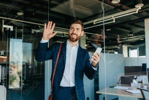 Happy male office worker in suit entering office space smiling and waving to colleagues, copy space