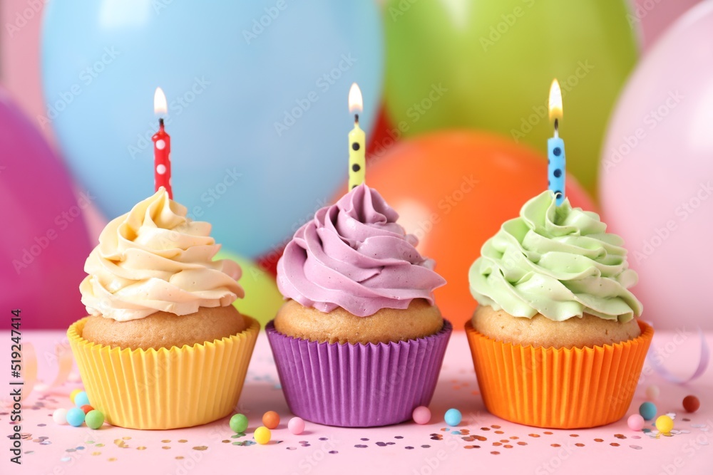 Birthday cupcakes with burning candles and sprinkles on pink table against color balloons, closeup Birthday cupcakes with burning candles and sprinkles on pink table against color balloons, closeup