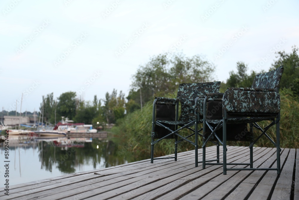 Fototapeta premium Camouflage fishing chairs on wooden pier near river, space for text