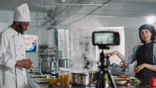 Multiethnic team of chefs hosting online cooking show, filming video on camera in restaurant kitchen. Man and woman doing gastronomic culinary class, recording food recipe preparations.