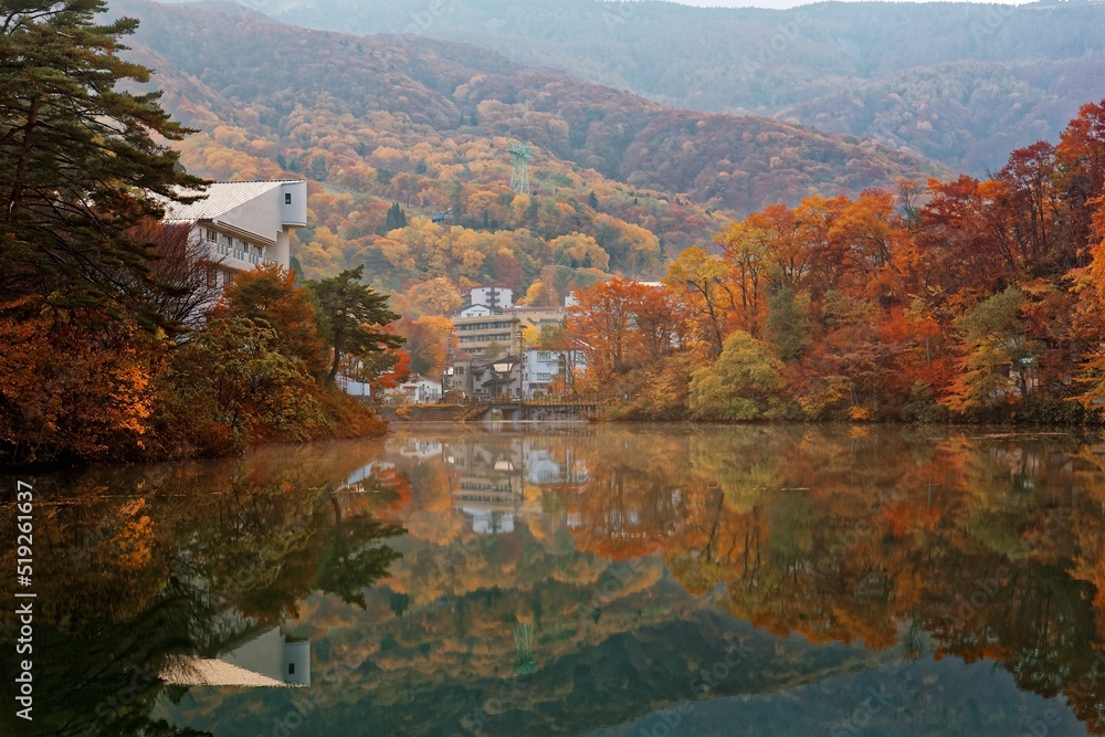 Fototapeta premium Scenery of an autumn pond with view of lakeside villas & houses in a peaceful atmosphere and fall foliage reflecting on the smooth water at Sakazuki Lake in Zao Onsen Resort in Yamagata, Japan