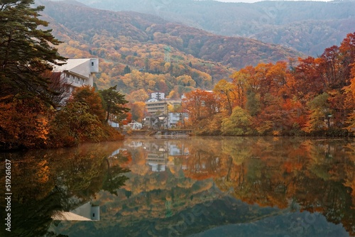 Scenery of an autumn pond with view of lakeside villas & houses in a peaceful atmosphere and fall foliage reflecting on the smooth water at Sakazuki Lake in Zao Onsen Resort in Yamagata, Japan