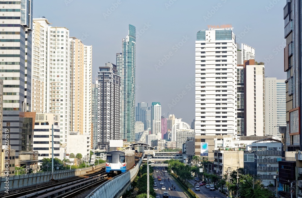 Fototapeta premium View of a BTS skytrain traveling on elevated metro system between high rise skyscrapers in Downtown Bangkok on a beautiful sunny day~ Cityscape of Bangkok, the fast developing capital city of Thailand