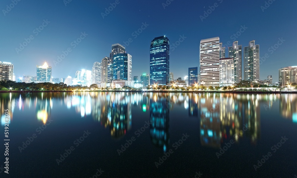 Naklejka premium Night skyline of modern lakeside skyscrapers with glass curtain walls and dazzling city lights reflected in the smooth lake water in beautiful Benjakiti Park at blue dusk, in Bangkok, Thailand, Asia