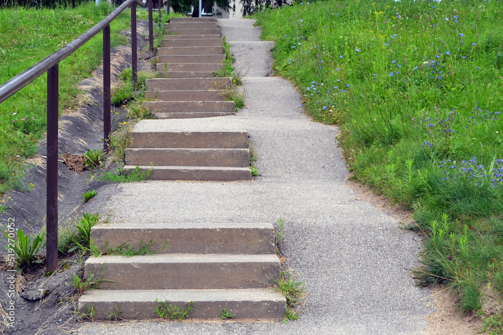 Stairs with gray concrete walkway and steps with railings lead up among green grass and flowers.
