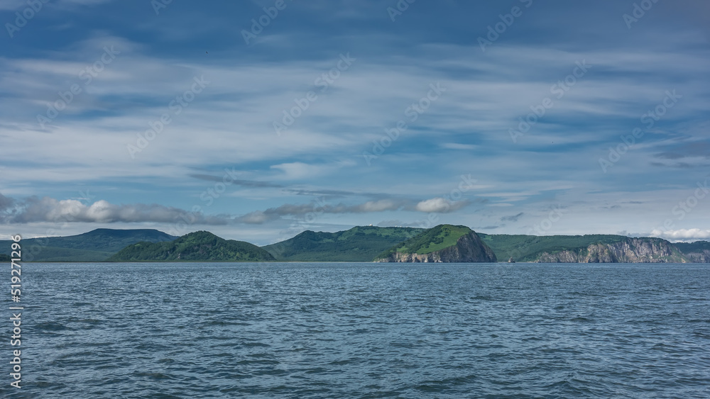 The picturesque coast of Kamchatka. Green hills and rocks against a background of blue sky, clouds and the Pacific Ocean. Avacha Bay