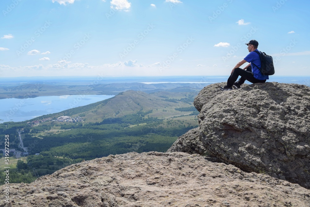 A mountaineer with a backpack sits on top of a cliff and looks at the lake on a summer day