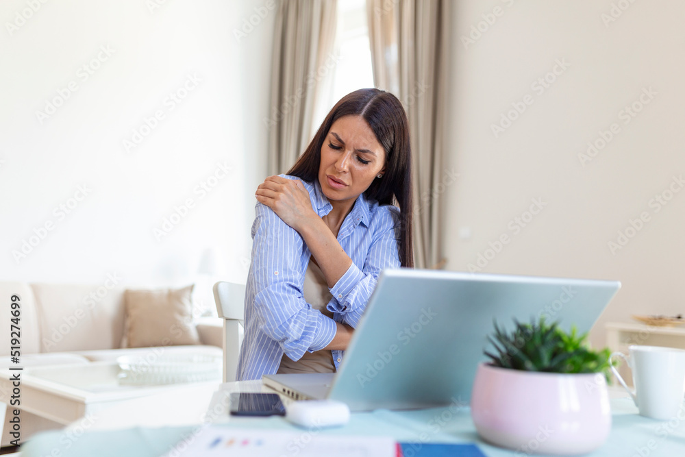 Portrait of young stressed woman sitting at home office desk in front of laptop, touching aching shoulder with pained expression, suffering from shoulder after working on laptop Portrait of young stressed woman sitting at home office desk in front of laptop, touching aching shoulder with pained expression, suffering from shoulder after working on laptop