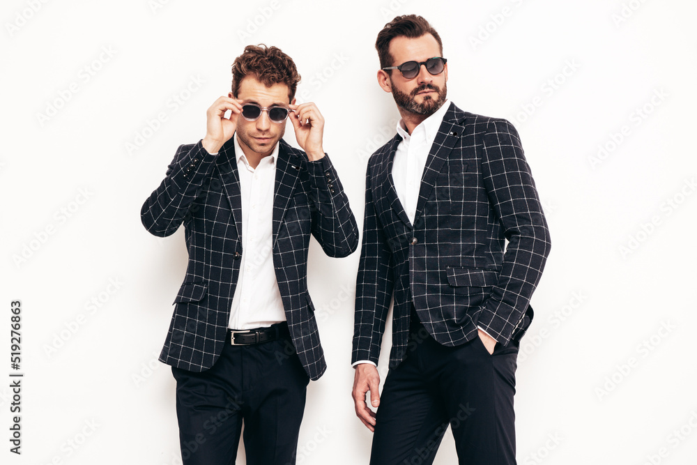 Portrait of two handsome confident stylish hipster lambersexual models. Sexy modern men dressed in black same elegant suit. Fashion male posing in studio near white wall. In sunglasses Portrait of two handsome confident stylish hipster lambersexual models. Sexy modern men dressed in black same elegant suit. Fashion male posing in studio near white wall. In sunglasses