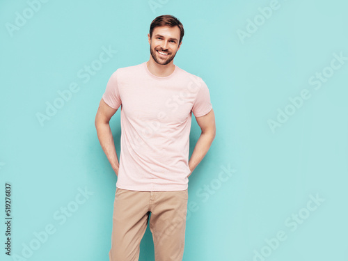Portrait of handsome smiling stylish hipster lambersexual model. Sexy man dressed in pink T-shirt and trousers. Fashion male isolated on blue wall in studio © halayalex