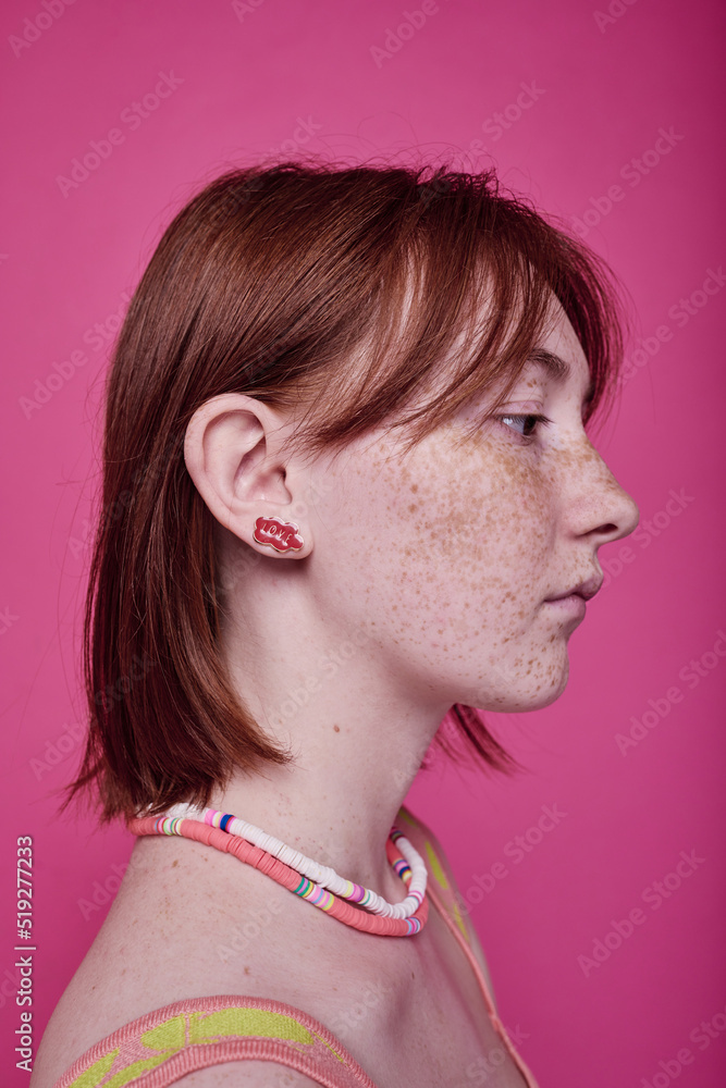 Side view of stylish teenage girl with short red hair with beads necklace on her neck posing at camera over pink background