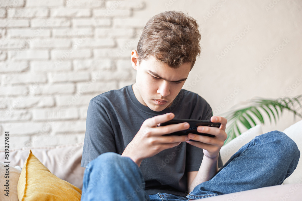 A teenage boy plays games on a smartphone with a focused expression. Dependence on the phone and online games. Close-up, bottom view. A young man with a phone in his hands sits on the couch. A teenage boy plays games on a smartphone with a focused expression. Dependence on the phone and online games. Close-up, bottom view. A young man with a phone in his hands sits on the couch.