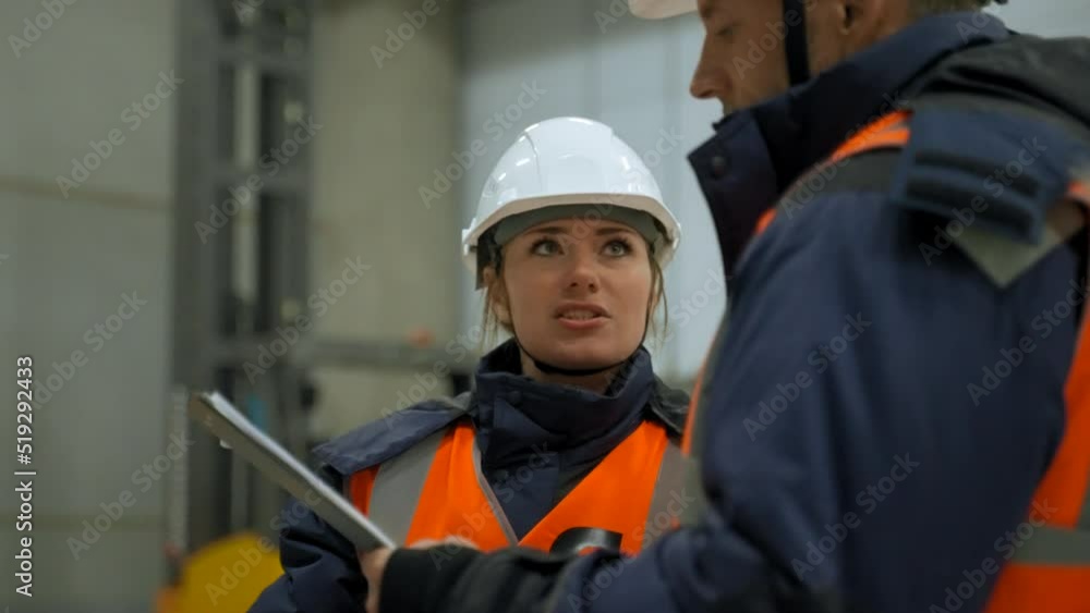 Woman and man in uniform discuss project plan looking at clipboard. Professional engineers in white helmets stand in warehouse closeup slow motion Woman and man in uniform discuss project plan looking at clipboard. Professional engineers in white helmets stand in warehouse closeup slow motion