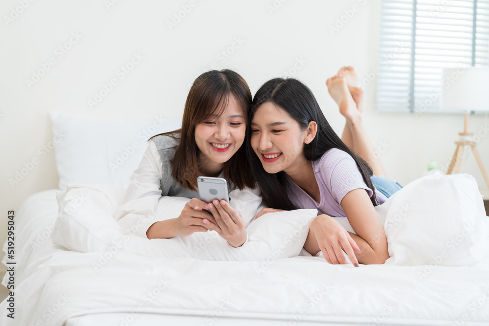 Asian beautiful two women use smartphone video call online with friend. Attractive young female couple lying down on bed in bedroom, holding mobile phone and talking virtual remote in morning in house Asian beautiful two women use smartphone video call online with friend. Attractive young female couple lying down on bed in bedroom, holding mobile phone and talking virtual remote in morning in house