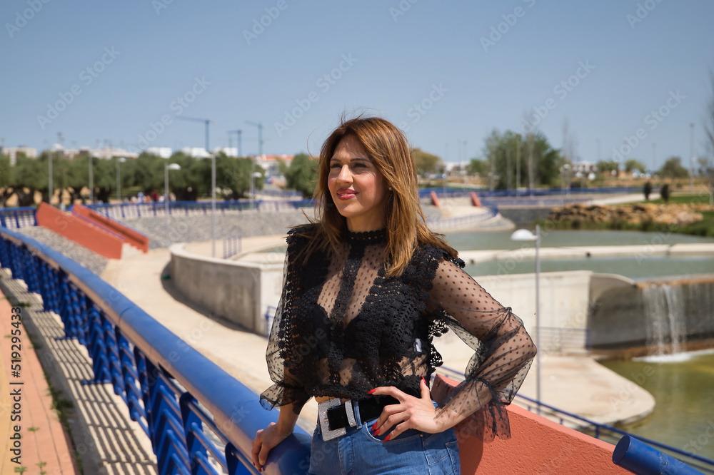Attractive mature woman in transparent black shirt and jeans, leaning on a railing in sensual and provocative attitude. Concept maturity, beauty, fashion, trend, sensuality, provocation.