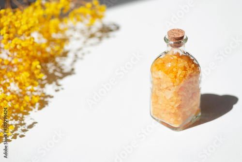 Transparent bottle filled with yellow bath sea salt with wild flowers on white background. Beauty treatment for spa and wellness. Skincare natural cosmetic concept for body care.